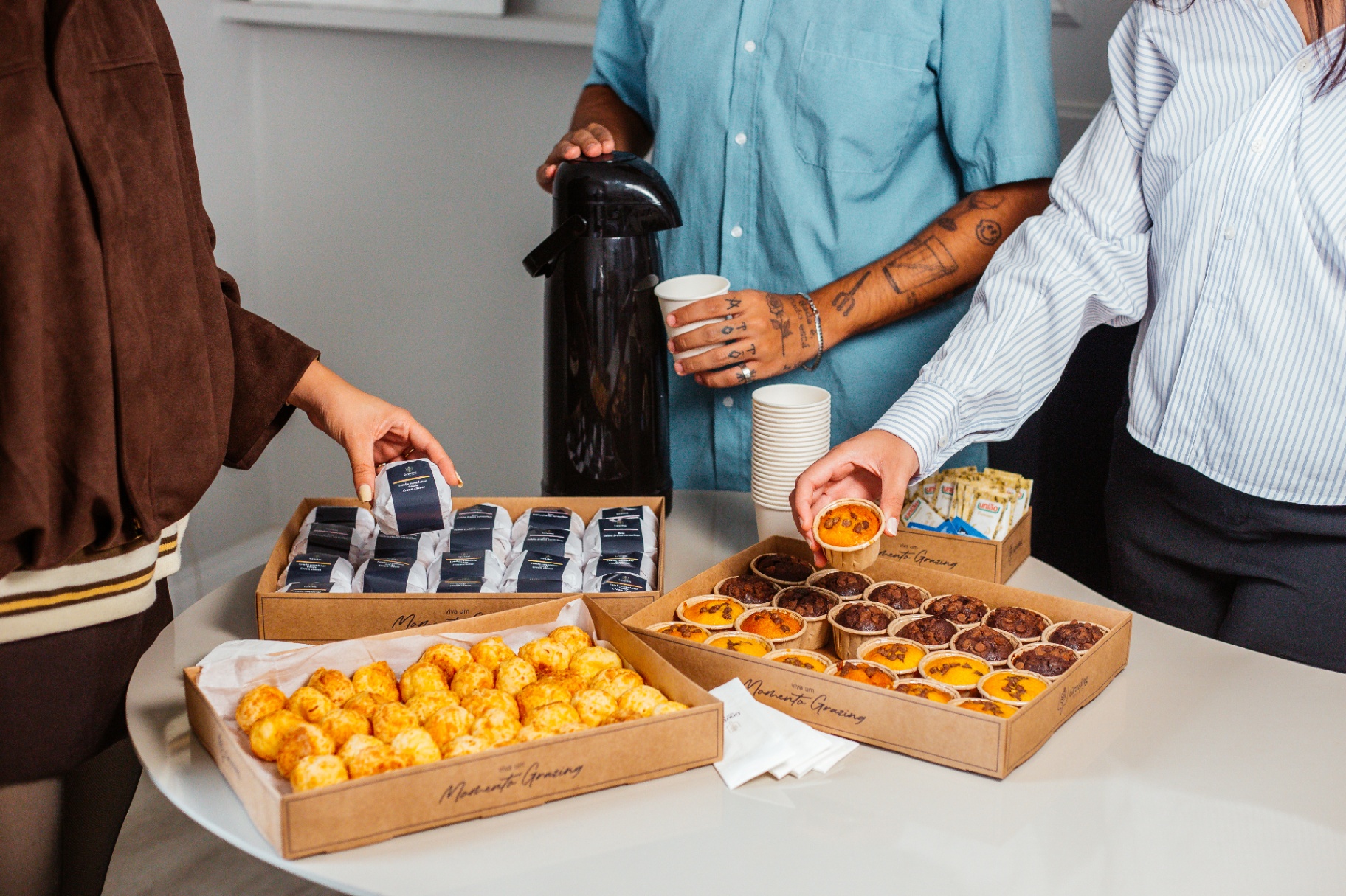Coffee break corporativo com caixas de salgados, doces e pessoas servindo-se